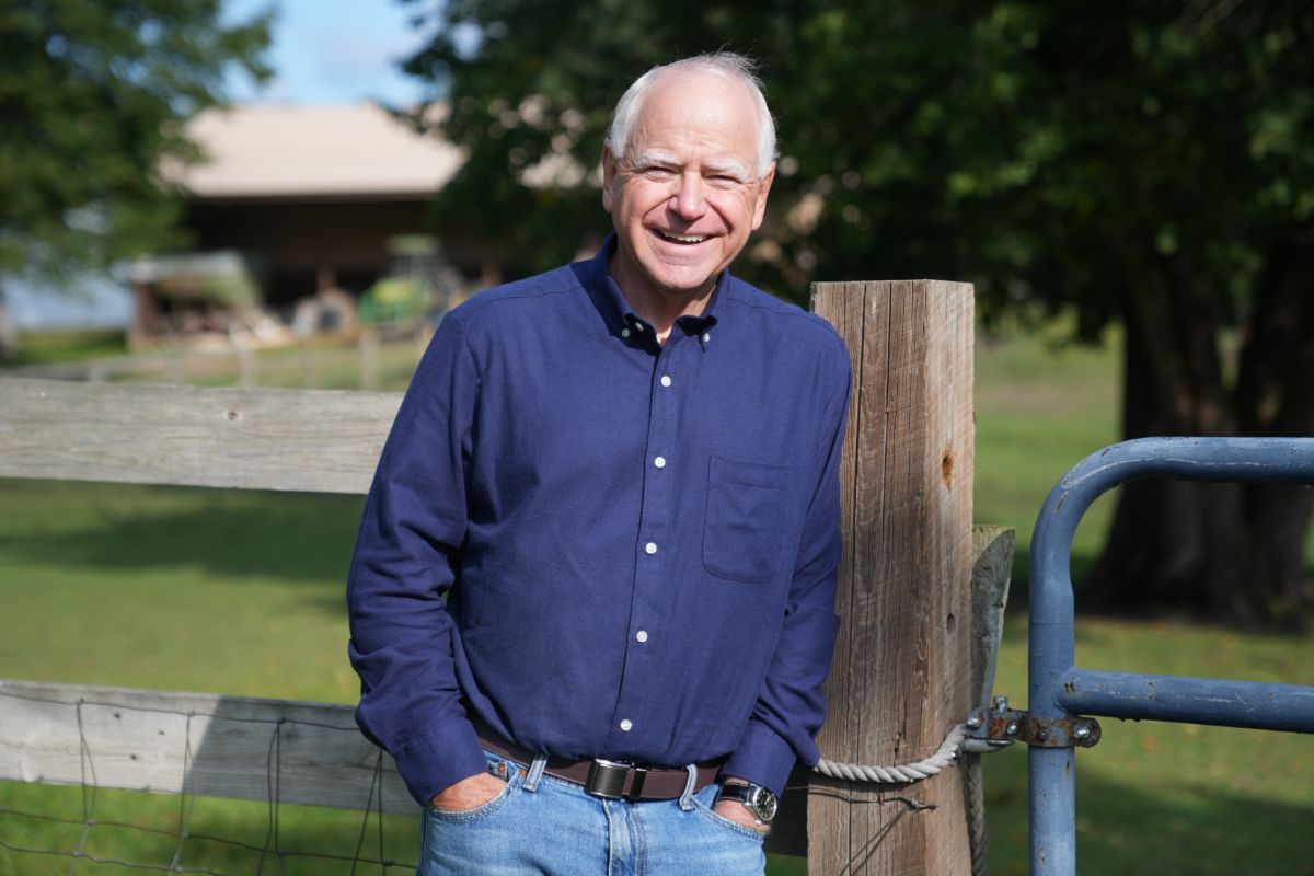 A picture of Tim Walz. He is wearing a blue shirt and standing in front of a fence gate.