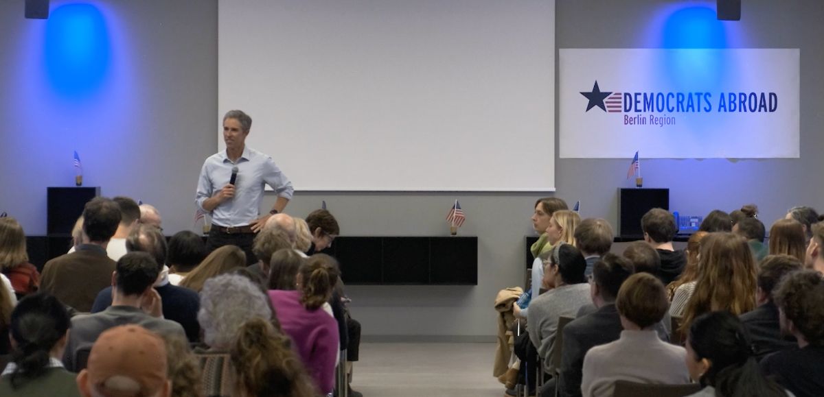 An image of Beto holding a microphone in front of a seated crowd. A sign on the wall behind reads 'Democrats Abroad: Berlin Region'.